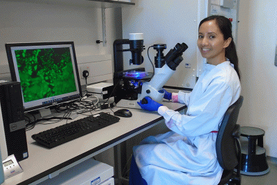 Amanda Corla in a lab coat and wearing purple gloves, sat on a chair in front of a microscope