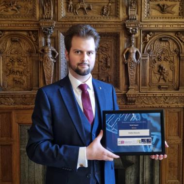 Stefan Dascalu in suit and tie, holds his Social Impact Award in front of wooden carved panel wall 