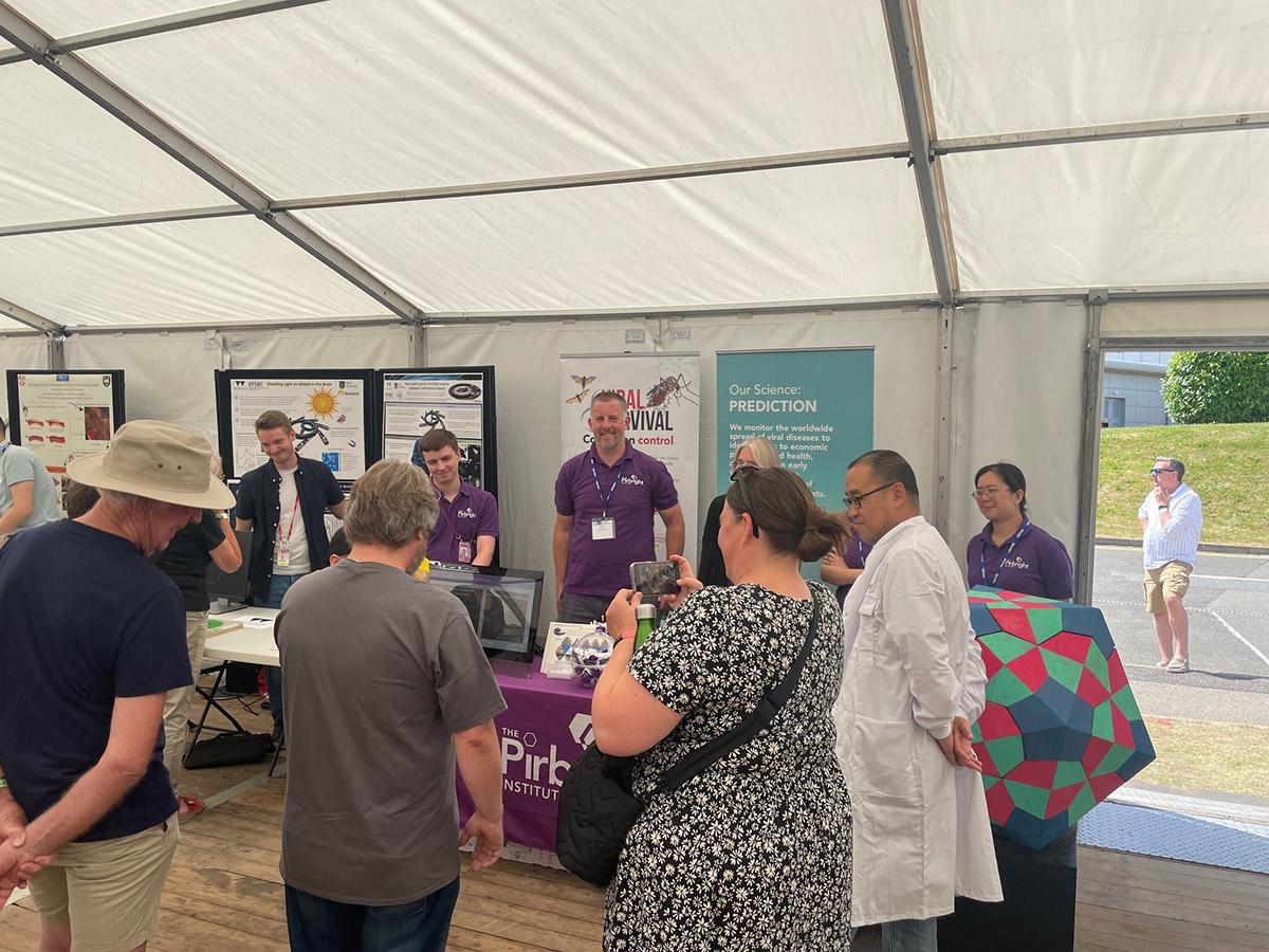 Family day at Harwell Group of Pirbright researchers in a marquee behind a table and in front of pull up posters with a small group of general public members looking at taking photos
