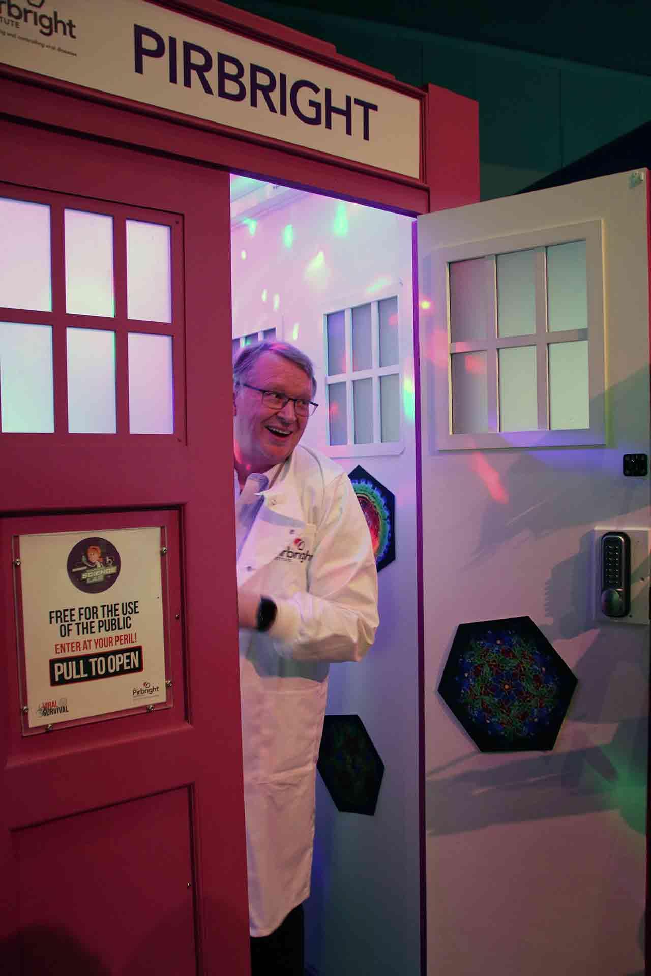 A smiling scientist in a white lab coat steps out of the door of the Dr Zoo's Viral Survival pink TARDIS